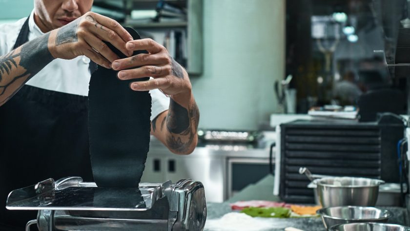Traditional italian food. Cropped image of chef's hands with tattoos rolling a black dough